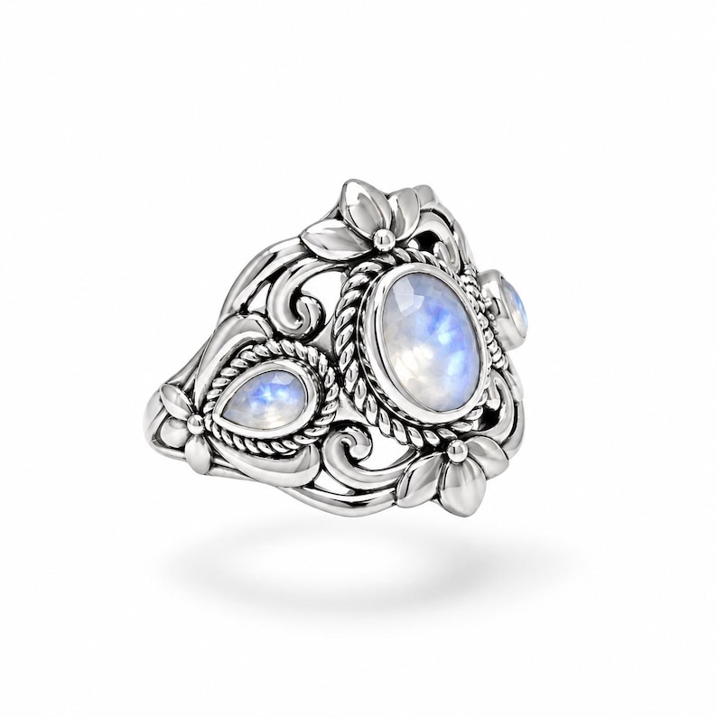 Puede incluir: Un anillo de plata con una piedra lunar ovalada grande y otra m&aacute;s peque&ntilde;a en forma de pera, engastadas en una banda de metal decorativa. La banda muestra intrincados adornos, detalles de cuerda trenzada y motivos florales alrededor de las piedras.