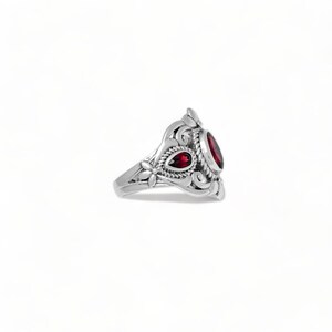 Puede incluir: Un anillo de plata con una piedra preciosa roja oscura de forma ovalada y una piedra roja m&aacute;s peque&ntilde;a en forma de l&aacute;grima. La banda de metal est&aacute; decorada con intrincados motivos de volutas, flores y una textura de cuerda.