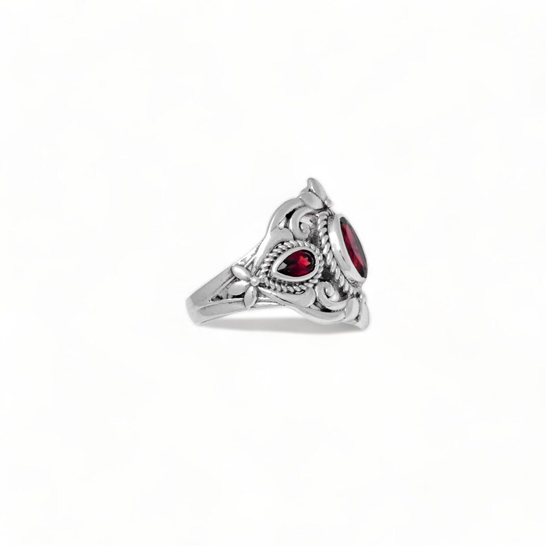 Puede incluir: Un anillo de plata con una piedra preciosa roja oscura de forma ovalada y una piedra roja m&aacute;s peque&ntilde;a en forma de l&aacute;grima. La banda de metal est&aacute; decorada con intrincados motivos de volutas, flores y una textura de cuerda.