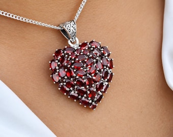 May include: A heart-shaped pendant necklace featuring numerous oval-cut deep red garnet gemstones set in silver. The pendant is attached to a silver chain, and the bail displays intricate engraved scrollwork details on its metallic surface.