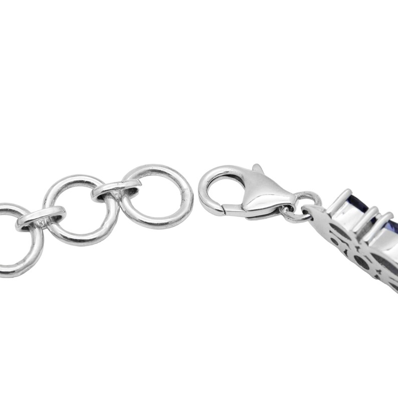 May include: Close-up of a silver bracelet with a lobster clasp and connecting rings. The bracelet features a decorative section with blue gemstones. The jewelry is set against a white background.