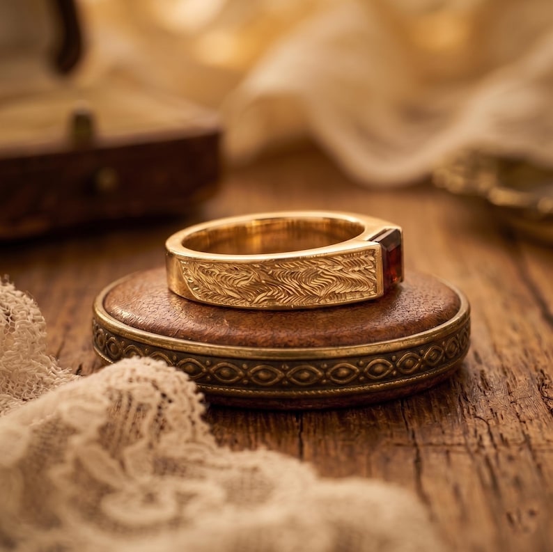 May include: A gold ring featuring a textured, wave-like pattern engraved along the band and a single, rectangular, dark red gemstone set at the front. The metal surface displays a polished finish with intricate detailing across the entire outer band.