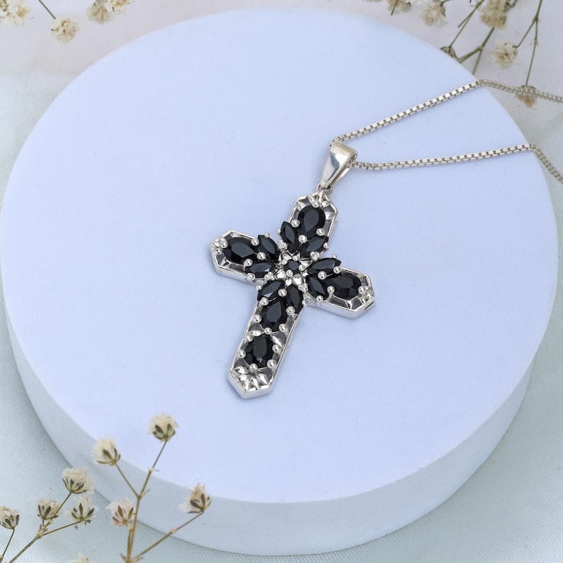 May include: A silver chain necklace with a black gemstone cross pendant. The cross is adorned with black gemstones in a floral pattern.