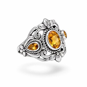 Puede incluir: Un anillo de plata con una gran citrina amarilla de corte ovalado en el centro, flanqueada por una citrina m&aacute;s peque&ntilde;a en forma de pera y una citrina redonda. La banda presenta intrincados motivos florales, volutas y detalles de cuerda.