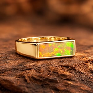 May include: A gold ring featuring a rectangular, horizontally set opal inlay. The opal displays a vibrant play of color with flashes of green, orange, and yellow. The band is polished gold with a smooth, solid finish and a flat, rectangular face.
