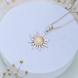 Op de afbeelding: Een zilveren ketting met een zon hanger met een witte opaal edelsteen. De hanger heeft een gedetailleerd zon ontwerp met stralen die vanuit het midden uitstralen.