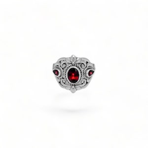 Puede incluir: Un anillo de plata con una piedra preciosa de color rojo intenso de corte ovalado en el centro, rodeada de intrincados adornos met&aacute;licos y detalles de cuerda. Dos piedras rojas m&aacute;s peque&ntilde;as en forma de l&aacute;grima flanquean la piedra central.