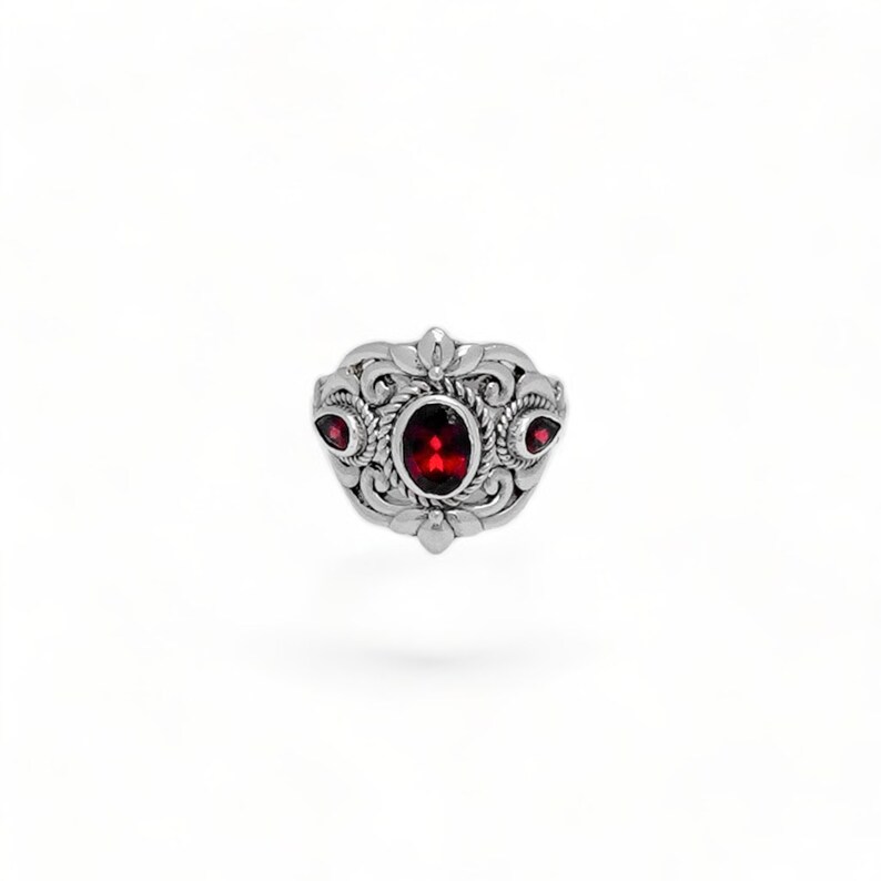 Puede incluir: Un anillo de plata con una piedra preciosa de color rojo intenso de corte ovalado en el centro, rodeada de intrincados adornos met&aacute;licos y detalles de cuerda. Dos piedras rojas m&aacute;s peque&ntilde;as en forma de l&aacute;grima flanquean la piedra central.