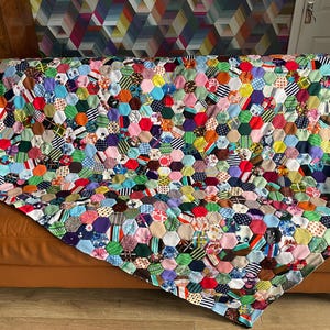 May include: A vibrant patchwork quilt with a honeycomb design, resting on a brown leather sofa. The quilt showcases a mix of fabrics in red, blue, green, and yellow hues. A geometric wallpaper forms the backdrop.
