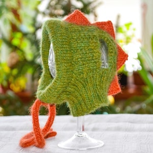 May include: A green and orange knitted dinosaur hat. The hat features a textured, green body with orange spikes along the head and orange ties. The hat is displayed on a clear glass.