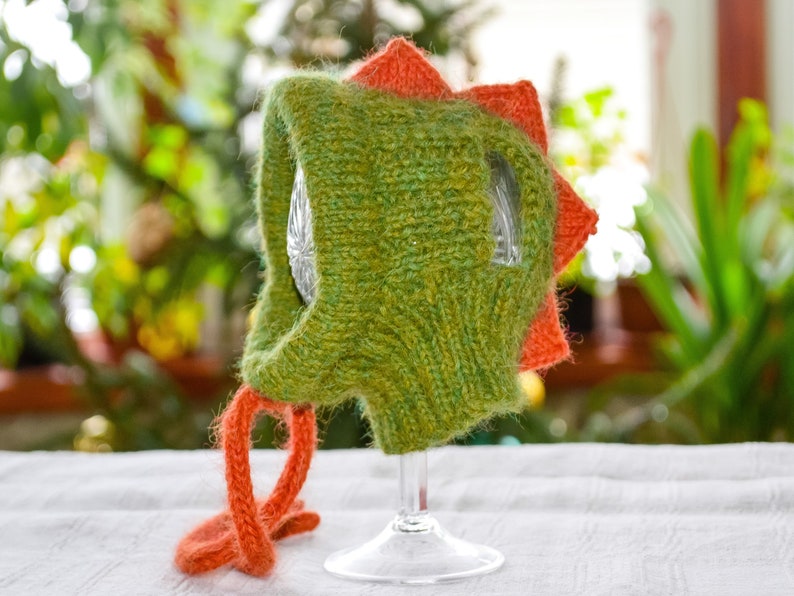 May include: A green and orange knitted dinosaur hat. The hat features a textured, green body with orange spikes along the head and orange ties. The hat is displayed on a clear glass.