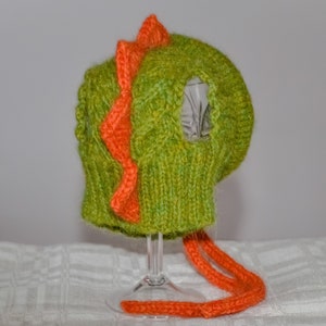 May include: A knitted green and orange dinosaur hat. The hat features a textured green knit with an orange zig-zag design down the center and orange ties. The hat is designed to cover the head and ears.