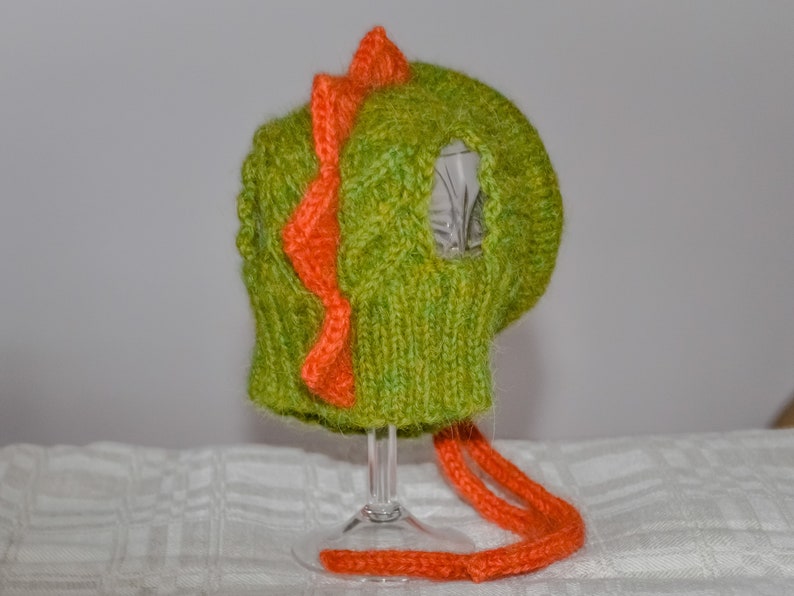 May include: A knitted green and orange dinosaur hat. The hat features a textured green knit with an orange zig-zag design down the center and orange ties. The hat is designed to cover the head and ears.