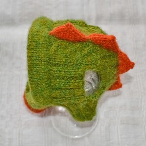 May include: A handmade green and orange knitted hat shaped like a dinosaur. The hat features a textured, fuzzy appearance with orange spikes along the top and a clear plastic eye. The hat is designed for children.