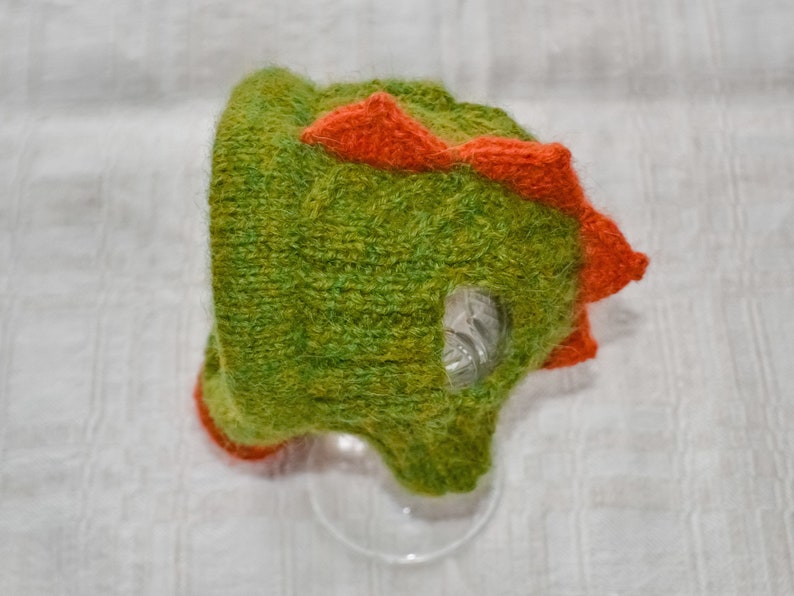 May include: A handmade green and orange knitted hat shaped like a dinosaur. The hat features a textured, fuzzy appearance with orange spikes along the top and a clear plastic eye. The hat is designed for children.