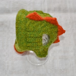 May include: A green and orange knitted dinosaur hat. The hat features a textured green body with orange spikes along the top and around the face opening. The hat is designed to resemble a dinosaur.