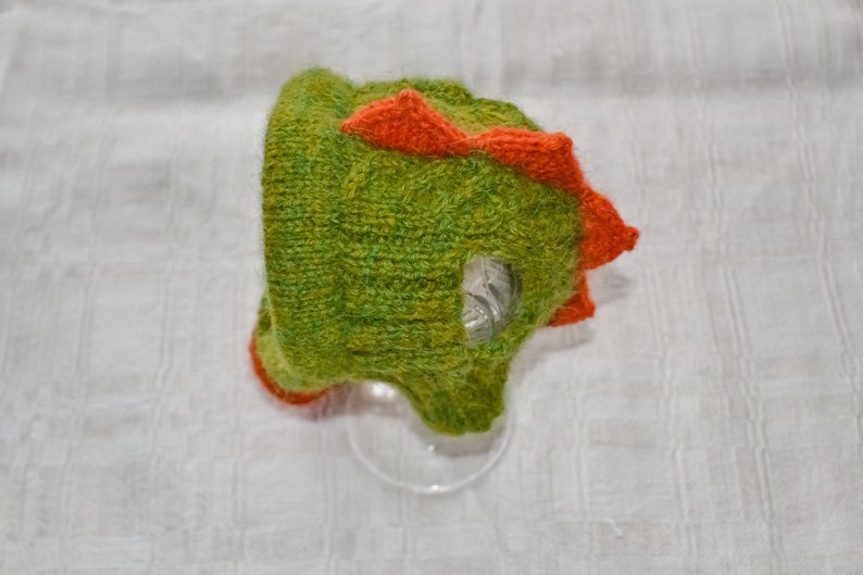 May include: A green and orange knitted dinosaur hat. The hat features a textured green body with orange spikes along the top and around the face opening. The hat is designed to resemble a dinosaur.
