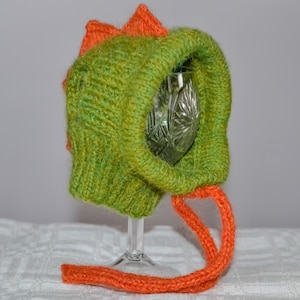 May include: A handmade green and orange knitted hat, resembling a dinosaur. The hat features a textured, ribbed knit design with an orange fin-like detail on top and an orange drawstring. The hat is designed to fit over a wine glass.