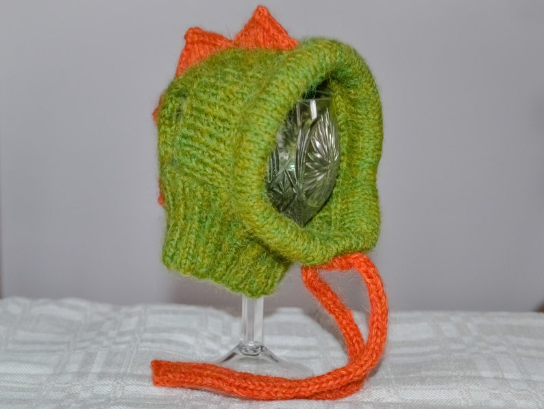 May include: A handmade green and orange knitted hat, resembling a dinosaur. The hat features a textured, ribbed knit design with an orange fin-like detail on top and an orange drawstring. The hat is designed to fit over a wine glass.