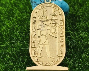 May include: Gold pendant necklace featuring an Egyptian god with a jackal head, standing with a staff, surrounded by hieroglyphs. The pendant is oval shaped and has a decorative base.