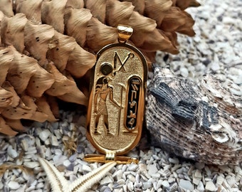 May include: Gold pendant featuring an Egyptian hieroglyphic design with a figure of a pharaoh holding a staff. The pendant is on a bed of small white stones and a seashell.