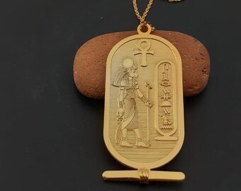 May include: A gold-tone necklace with a pendant featuring an Egyptian hieroglyphic design. The pendant is oval-shaped and depicts a figure standing with a staff, surrounded by hieroglyphs. The necklace is made of a delicate chain.