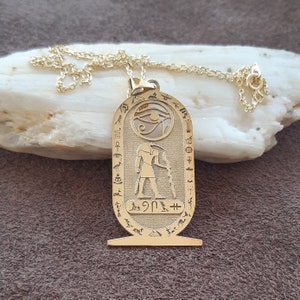 Könnte beinhalten: Goldene Halskette mit einem ägyptischen Horus-Augen-Anhänger. Der Anhänger zeigt eine detaillierte Darstellung des Horus-Auges, ein Symbol für Schutz und Gesundheit, umgeben von Hieroglyphen. Die Halskette ist aus Gold und hat einen Gliederketten-Stil.