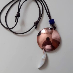 Può includere: Una collana con cordino marrone scuro dotato di nodi regolabili, un ciondolo circolare in rame lucido, una piccola perla cubica blu e un ciondolo bianco ricurvo a forma di dente appeso alla parte inferiore del disco in rame.