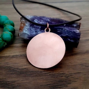 Puede incluir: Un colgante de color cobre con un diseño circular cuelga de un cordón negro. El colgante descansa sobre una piedra azul oscuro y marrón, con una pulsera de cuentas turquesa en el fondo. Una pieza de joyería sencilla y elegante.