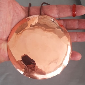 May include: A circular, polished copper disc with a reflective surface, suspended by two brown cords. The disc is held in a hand, and a red ring is visible on a finger. The copper has a warm, reddish-brown hue.