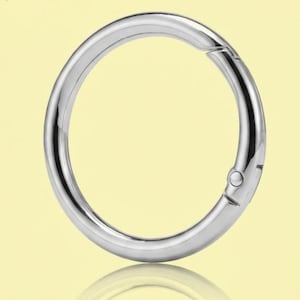 May include: A polished silver-tone metal spring gate ring with a circular shape. The hardware features a hinged mechanism on one side that allows the ring to open and close securely, showcasing a smooth, reflective surface and a clean, rounded profile.