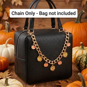 Op de afbeelding: Een goudkleurige metalen ketting met kleine bedeltjes in de vorm van oranje pompoenen en gele zonnebloemen. De ketting hangt over een zwarte handtas met een textuur. De tekst "Chain Only - Bag not included" staat op de afbeelding.