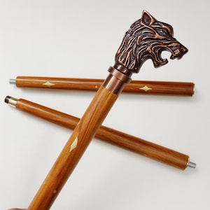 May include: A wooden walking stick with a bronze wolf head handle. The stick has a light brown finish with diamond-shaped inlays. The wolf head is detailed with textured fur and an open mouth. The stick is approximately 91 cm long.