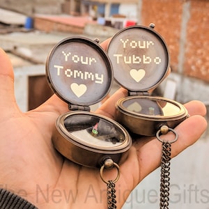 Op de afbeelding: Twee open antieke messing kompassen, elk gegraveerd met "Your Tommy" en "Your Tubbo" boven een hartsymbool. De kompassen hebben een ketting en worden in een hand gehouden tegen een zonsondergang.