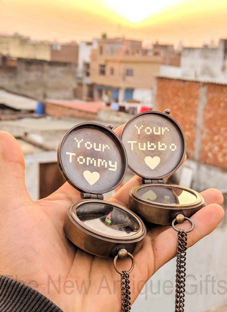 K&ouml;nnte beinhalten: Zwei offene antike Messingkompasse, jeweils mit "Your Tommy" und "Your Tubbo" &uuml;ber einem Herzsymbol graviert. Die Kompasse haben eine Kette und werden in einer Hand vor einem Sonnenuntergangshintergrund gehalten.