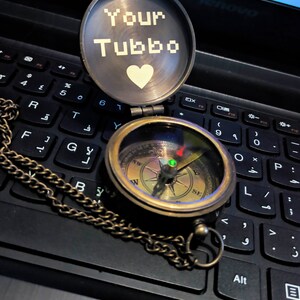 K&ouml;nnte beinhalten: Ein Kompass im Vintage-Stil aus Messing mit Kette, ge&ouml;ffnet auf einer Laptop-Tastatur. Das Kompasszifferblatt ist sichtbar, und der Deckel zeigt den Text "Your Tubbo" &uuml;ber einem wei&szlig;en Herz. Ein klassisches Navigationswerkzeug.