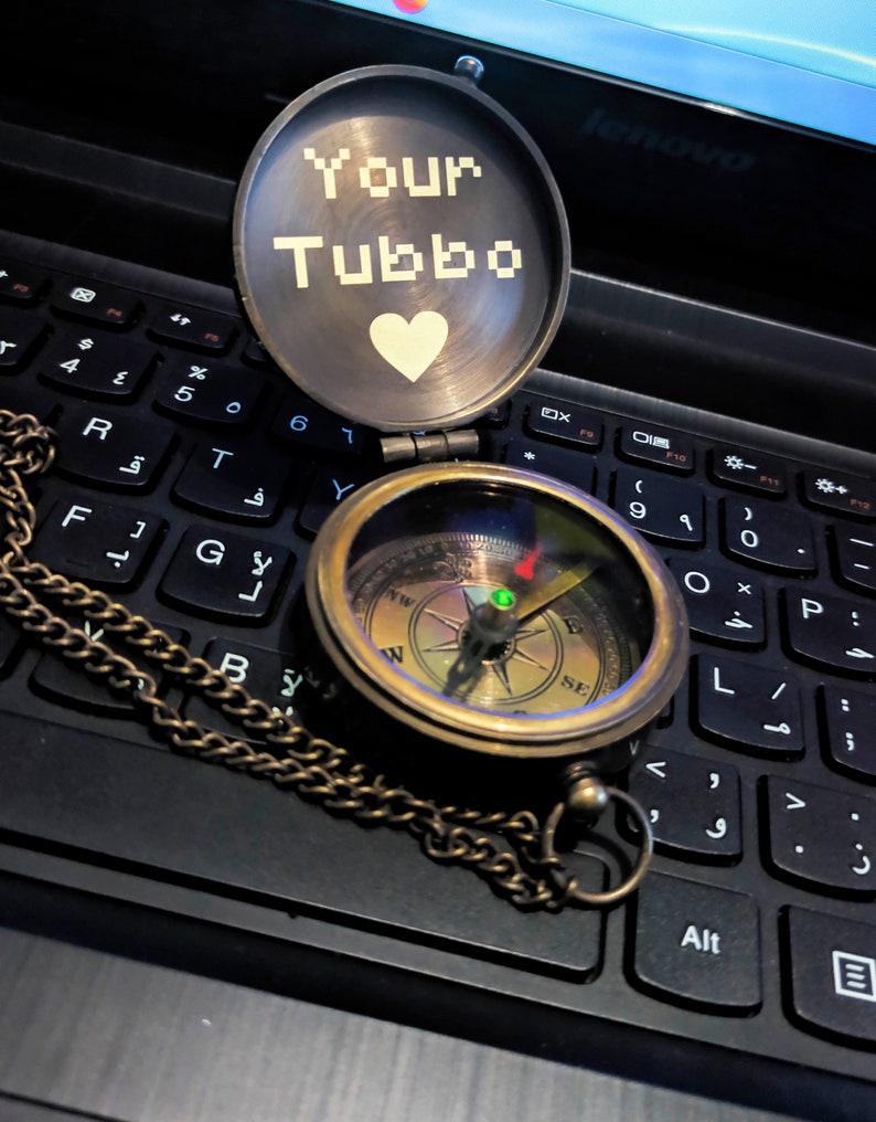 K&ouml;nnte beinhalten: Ein Kompass im Vintage-Stil aus Messing mit Kette, ge&ouml;ffnet auf einer Laptop-Tastatur. Das Kompasszifferblatt ist sichtbar, und der Deckel zeigt den Text "Your Tubbo" &uuml;ber einem wei&szlig;en Herz. Ein klassisches Navigationswerkzeug.