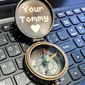 K&ouml;nnte beinhalten: Ein offener antiker Messingkompass mit Kette, der auf einer Laptop-Tastatur liegt. Das Kompasszifferblatt zeigt Richtungsmarkierungen. Der Deckel tr&auml;gt die Aufschrift "Your Tommy" mit einem Herzsymbol. Die Kompassnadel zeigt nach Osten.
