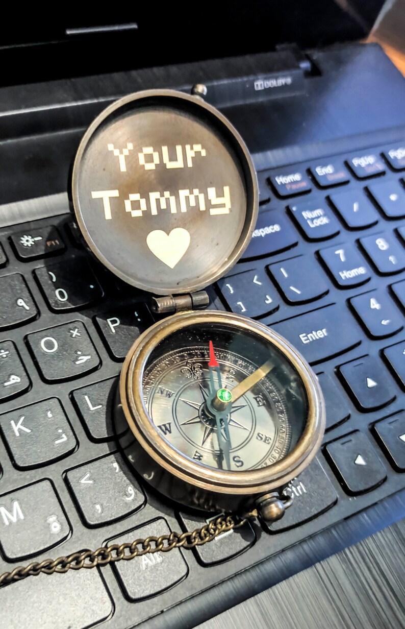 K&ouml;nnte beinhalten: Ein offener antiker Messingkompass mit Kette, der auf einer Laptop-Tastatur liegt. Das Kompasszifferblatt zeigt Richtungsmarkierungen. Der Deckel tr&auml;gt die Aufschrift "Your Tommy" mit einem Herzsymbol. Die Kompassnadel zeigt nach Osten.