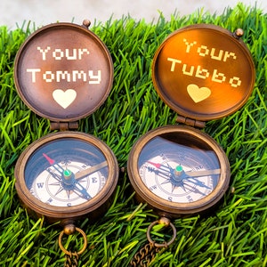 K&ouml;nnte beinhalten: Zwei antike Messingkompasse mit den Texten "Your Tommy" und "Your Tubbo" auf den Deckeln, jeweils mit einem Herzsymbol. Die Kompasse mit Ketten liegen auf gr&uuml;nem Gras.