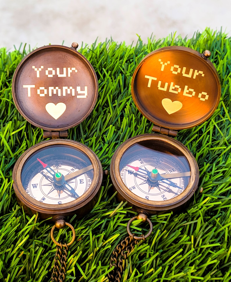 K&ouml;nnte beinhalten: Zwei antike Messingkompasse mit den Texten "Your Tommy" und "Your Tubbo" auf den Deckeln, jeweils mit einem Herzsymbol. Die Kompasse mit Ketten liegen auf gr&uuml;nem Gras.