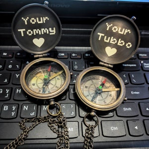 K&ouml;nnte beinhalten: Zwei antike Messingkompasse mit Ketten, ge&ouml;ffnet, um den Text "Your Tommy" und "Your Tubbo" mit einem Herzsymbol zu zeigen. Die Kompasse haben ein detailliertes Zifferblatt mit Richtungsmarkierungen und einer Nadel. Die Kompasse liegen auf einer schwarzen Tastatur.