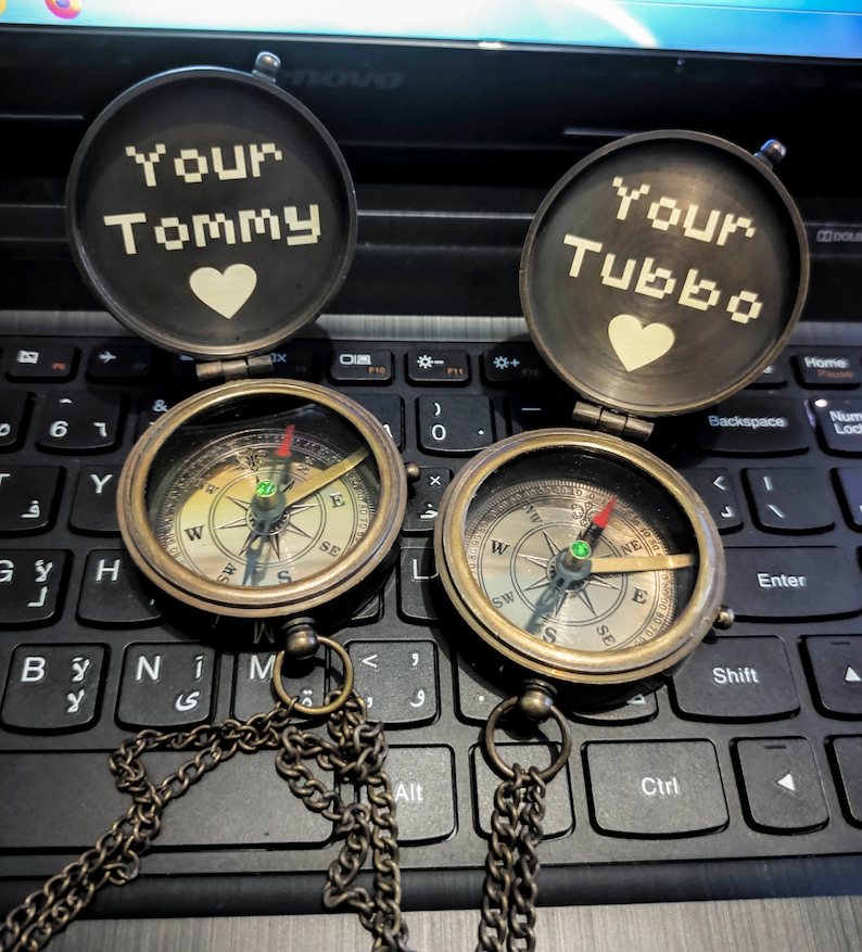 K&ouml;nnte beinhalten: Zwei antike Messingkompasse mit Ketten, ge&ouml;ffnet, um den Text "Your Tommy" und "Your Tubbo" mit einem Herzsymbol zu zeigen. Die Kompasse haben ein detailliertes Zifferblatt mit Richtungsmarkierungen und einer Nadel. Die Kompasse liegen auf einer schwarzen Tastatur.