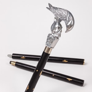 Puede incluir: Un bastón negro con un diseño de pájaro y calavera plateados. El bastón tiene detalles dorados y está sobre una superficie blanca. El pájaro está posado sobre una calavera, añadiendo un elemento decorativo único.
