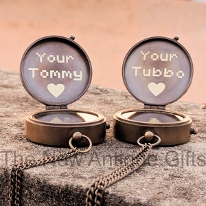 K&ouml;nnte beinhalten: Zwei antike Messingkompasse mit offenen Deckeln, jeweils mit "Your Tommy" und "Your Tubbo" &uuml;ber einem Herzsymbol graviert. Die Kompasse haben Ketten und liegen auf einer Steinoberfl&auml;che. Der Text "The New Antique Gifts" befindet sich unten.