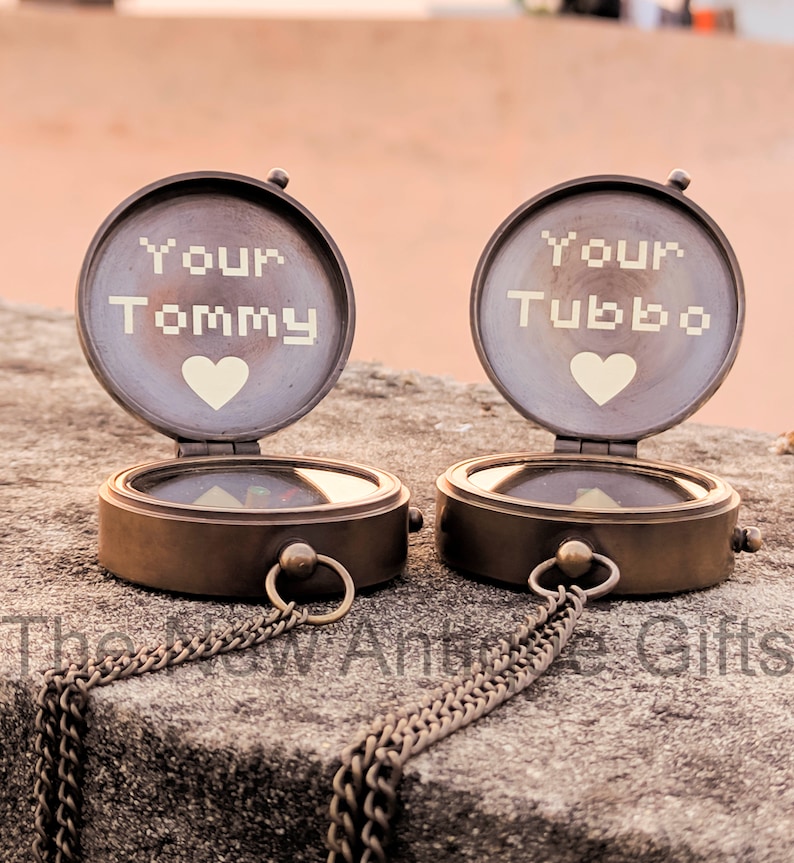 K&ouml;nnte beinhalten: Zwei antike Messingkompasse mit offenen Deckeln, jeweils mit "Your Tommy" und "Your Tubbo" &uuml;ber einem Herzsymbol graviert. Die Kompasse haben Ketten und liegen auf einer Steinoberfl&auml;che. Der Text "The New Antique Gifts" befindet sich unten.