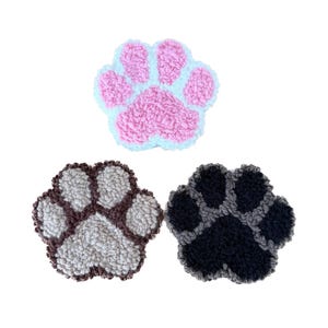 Könnte beinhalten: Drei plüschige, pfotenförmige Artikel in Rosa, Braun und Schwarz. Die rosa Pfote hat eine hellblaue Umrandung. Die braune Pfote hat eine hellgraue Mitte. Die schwarze Pfote hat eine dunkelgraue Mitte. Alle drei haben ein strukturiertes, geschlungenes Aussehen.