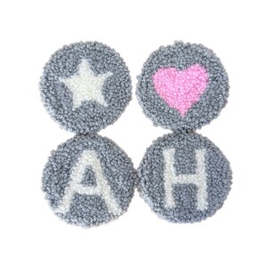 Könnte beinhalten: Vier graue, kreisförmige, getuftete Untersetzer. Einer zeigt einen weißen Stern, ein anderer ein rosa Herz, und die anderen zeigen die weißen Buchstaben "A" und "H". Die Untersetzer sind in einem 2x2-Raster angeordnet.