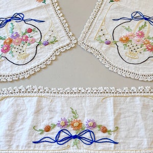 May include: Three white crocheted tablecloths with embroidered floral designs. The center tablecloth features a blue bow and a floral arrangement. The two side tablecloths feature a basket of flowers with a blue bow.