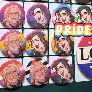 May include: A selection of round pin badges with illustrations of two characters with long hair and beards. Some badges display Pride flags, and one has the word "PRIDE" in rainbow letters. Another badge has the word "LOVE" on it.